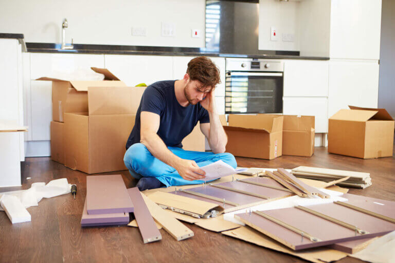 En man som sitter på golvet och monterar möbler med instruktioner, omgiven av kartonger i ett kök.